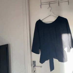 Svart omlottopp med knyt i midjan - Säljer en svart omlottopp med breda trekvartsärmar och snygg knytning i ryggen. Toppen har en rundad halsringning och är tillverkad i ett mjukt tyg som ger en skön känsla. Perfekt för dig som gillar stilrena och trendiga plagg med lite extra detalj.