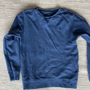 Blå tröja från Polo Ralph Lauren - Säljer en klassisk  blå college tröja från Polo Ralph Lauren i lite tunnare material med rund hals och diskret broderad logga på bröstet. Tröjan har lång ärm och är tillverkad i mjuk bomull, perfekt för en stilren och avslappnad look. Passar pojke 12-16 år eller liten man,xs. Lite blekt men inget som stör