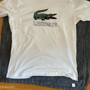 Vit Lacoste t-shirt med krokodiltryck - Vit t-shirt från Lacoste med klassiskt krokodiltryck och logga på bröstet. Modellen är kortärmad och har rund hals. Materialet är mjuk bomull som känns skönt mot huden. Perfekt för dig som gillar stilrena och ikoniska plagg.