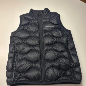 Svart dunväst Peak Performance - Svart dunväst från Peak Performance med quiltad design och hög krage. Västen har två sidofickor med dragkedja och logotyp broderad på bröstet. Materialet är glansigt och känns lätt, perfekt för lager-på-lager. Snygg och sportig look. Ny pris 1299kr