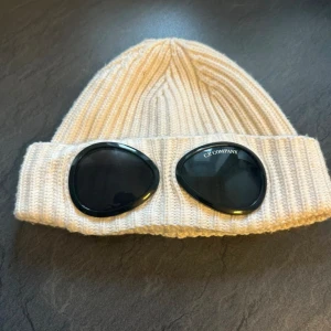 Vit C.P. Company mössa - Säljer en mössa från C.P. Company med svarta goggle-linser framtill. Mössan är tillverkad i mjukt stickat material och har en bred uppvikt kant med de klassiska glasögon-detaljerna. Perfekt till dig som behöver en snygg mössa i vinter ny pris 1299kr