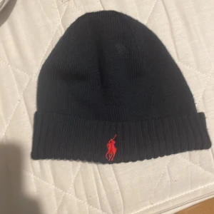 Mörkblå mössa från Polo Ralph Lauren - Snygg mörkblå mössa från Polo Ralph Lauren i mjuk merinoull. Klassisk ribbstickad design med uppvikt kant och röd broderad Polo-logga framtill. Perfekt accessoar för kalla dagar och enkel att matcha med olika outfits.