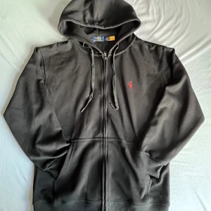 Svart hoodie från Polo Ralph Lauren - Svart hoodie från Polo Ralph Lauren med dragkedja och huva. Klassisk röd broderad logga på bröstet och snörning i huvan. Mjukt material och känguruficka framtill. Perfekt för en chill och stilren look.