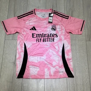 Rosa Real Madrid fotbollströja Adidas - Snygg rosa Real Madrid fotbollströja från Adidas med svart logga och klubbmärke på bröstet. Tröjan har korta ärmar, svarta detaljer och ett unikt mönster i olika rosa nyanser. Tillverkad i lätt och ventilerande polyester med Aeroready-teknologi.