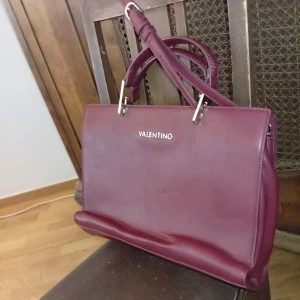 Vinröd handväska från Valentino - Snygg vinröd handväska från Valentino i skinn med stilrena detaljer och både handtag och axelrem. Väskan har en klassisk rektangulär form och insidan är fodrad med ljusbeige tyg med Valentino-logga. Perfekt för dig som gillar lyxiga accessoarer.