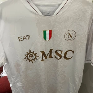 Vit Napoli fotbollströja EA7 - Snygg vit fotbollströja från Napoli med gulddetaljer och diskret mönster i tyget. Tröjan har klubbmärke, italienska mästarmärket och MSC-logga på bröstet. Priset går att diskutera vid snabb affär 
