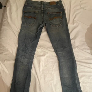 Blå jeans från Nudie Jeans - Säljer ett par klassiska blå jeans från Nudie Jeans med orangea sömmar och signaturbroderi på bakfickorna. Jeansen har en rak passform och är tillverkade i slitstarkt bomullsdenim. Perfekta för dig som gillar en tidlös och avslappnad stil.