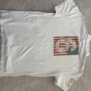 Polo Jeans Company by Ralph Lauren – Klassisk vit piké (strl L) - Klassisk vit piké från Polo Jeans Company by Ralph Lauren, storlek L. Äkta vintage-plagg från sent 90-/tidigt 2000-tal – sällsynt och eftertraktad modell! Broderad logga på bröstet, stort retrotryck med amerikanska flaggan på ryggen och mjukt, högkvalitativt tyg.  ✨ Skick: Mycket bra – endast lätt använd, inga fläckar eller hål. 📏 Storlek: L (normal passform) 📦 Skickas snabbt och välpackat!  💰 Pris: 299 kr (värd betydligt mer som vintage Ralph Lauren!)