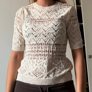 Stickad vit topp med kort ärm - Säljer en superfin stickad topp i vit färg med korta ärmar och snyggt hålmönster. Tröjan har rund halsringning och ett detaljerat zigzag-mönster som ger en luftig och trendig look. Perfekt för vår och sommar när du vill ha något lätt och snyggt.