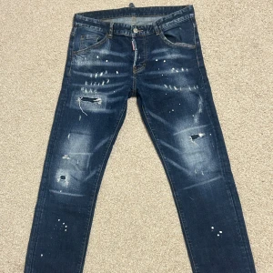 Dsquared2 blå distressed jeans, stl 46 - Snygga blå jeans från Dsquared2 i storlek 46 med coola slitningar, färgstänk och distressed detaljer. Klassisk femficksmodell med raka ben och tydliga slitningar framtill och baktill. Perfekta för dig som gillar en edgy streetstil.