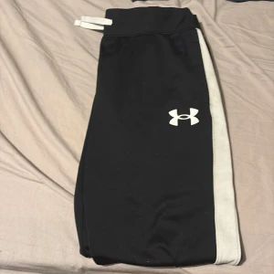 Svarta mjukisbyxor från Under Armour - Svarta mjukisbyxor från Under Armour med vita sidor och logga på låret. Byxorna har dragsko i midjan och är tillverkade i ett mjukt syntetmaterial som passar perfekt för träning eller chill. Snygg och sportig look med smal passform.