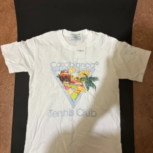 Casablanca Tennis Club t-shirt - Vit t-shirt från Casablanca med färgglatt tryck framtill och texten 'Casablanca Tennis Club'. Klassisk rund hals och korta ärmar. Skön bomullskvalitet och avslappnad passform, perfekt för en sportig och trendig look.