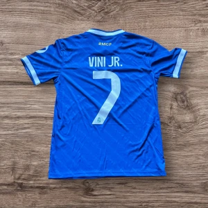 Real Madrid Vini Jr. blå matchtröja S - Blå Real Madrid fotbollströja med Vini Jr. och nummer 7 på ryggen. Adidas-logga och klubbmärke på bröstet, samt vita detaljer på krage och ärmslut. Kortärmad modell i lätt och ventilerande polyester, med tryckta sponsors och Aeroready-teknologi.