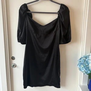 Svart kort klänning från Parisian - Snygg svart kort klänning från Parisian med puffiga korta ärmar och v-ringad hals. Klänningen har en figurnära passform och dragkedja bak. Materialet är glansigt och känns mjukt mot huden, perfekt för dig som gillar stilrena och trendiga plagg. Endast testad, aldrig använd 