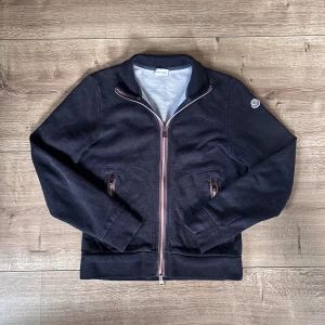 Grå cardigan tröja från Moncler - Mörk grå cardigan tröja från Moncler med ribbade muddar, ståkrage och hel dragkedja med röd, vit rand. Tröjan har två fickor med dragkedja och Moncler-logga på vänster ärm. Mjuk bomullskänsla och stilren look. Storleken är M men den är liten i storlek så den sitter som en S. Dragkedjan har också gått sönder där nere vid knäppet men man lär sig snabbt att stänga den då den kan va lite svår i början att stänga. -därav pris men priset går att diskutera vid snabb affär med!