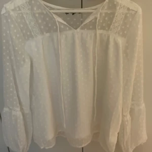 Vit prickig blus med spetsdetaljer - Supersöt vit blus med prickigt mönster och transparenta partier. Blusen har långa ärmar med volang och spetsdetaljer samt knytband vid halsen. Perfekt för dig som gillar romantisk och luftig stil.