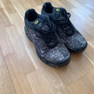 Nike Air Max Plus svarta sneakers - Nike Air Max Plus sneakers med svart och grått mönstrat tyg, svarta detaljer och snörning. Snygg streetstil med robust sula och Tuned Air-dämpning. Ikonisk TN-logga på plösen och hälen. Perfekt för dig som gillar unika sneakers med attityd.