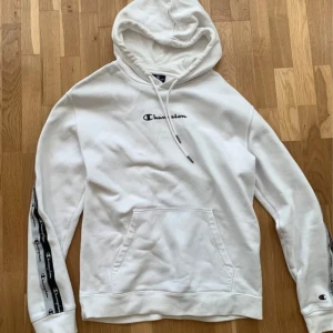 Vit hoodie från Champion med logga - Vit hoodie från Champion med svart broderad logga på bröstet och loggoband längs ärmarna. Klassisk känguruficka framtill, huva med snörning och ribbade muddar. Perfekt för en chill och sportig look.