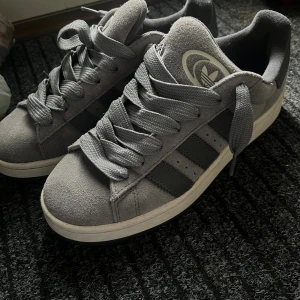 Grå Adidas sneakers i mocka - Snygga grå sneakers från Adidas med klassiska svarta ränder på sidorna och logga på plösen. Skorna har breda grå snören och en mjuk mocka-överdel. Yttersulan är vit med svart undersida och har en platt sula. Perfekt för dig som gillar stilrena och sportiga skor.