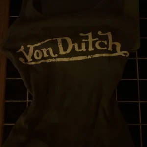 ribbad linnetopp Von Dutch - Vintage kakigrön ribbad linnetopp från Von Dutch med stort vitt logotryck framtill. Toppen har en tight passform och är ärmlös, perfekt för att ge en retro vibe till din outfit. Snygg att styla med jeans eller kjol för en trendig look.