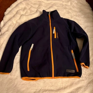 Mörkblå fleecejacka från Stormberg. 7år - Mörkblå fleecejacka från Stormberg med orange dragkedjor och detaljer. Jackan har hög krage, tre fickor med dragkedja och reflexdetaljer baktill och på blixtlåsen . Perfekt för kyliga dagar och outdoor-häng. Broderad logga på bröstet och ryggen.