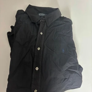 Svart skjorta från Polo Ralph Lauren - Klassisk svart skjorta från Polo Ralph Lauren med blå broderad logga på bröstet. Skjortan har vita knappar, diskreta ränder i tyget och kontrastsömmar. Tillverkad i 100% bomull och har lång ärm med manschettknappar.