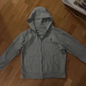 Grå hoodie från Polo Ralph Lauren - Grå hoodie från Polo Ralph Lauren med dragkedja och huva. Klassisk design med broderad logga på bröstet och två fickor framtill. Tillverkad i mjuk bomullsmix, perfekt för chill dagar eller när du vill ha en clean look.