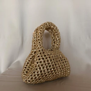 Gullig handväska -  En virkad liten väska gjord i ett tjockt guldigt garn! Väskan är 20cm bred och 9cm djup, höjden för väskan mätt med handtag är 25cm och 14cm när jag har måttat nedanför  handtagen. 