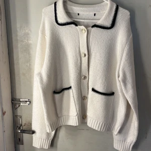 Kofta - Superfin vit kofta med svarta detaljer vid krage och fickor. Guldiga knappar framtill och två fickor. Koftan är stickad och har en klassisk, lite boxig passform med långa ärmar och ribbade muddar. Perfekt för lager-på-lager under kyliga dagar.