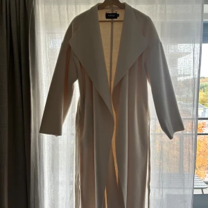 Vit kappa Bianca x Nelly.com  - Benvit kappa från Bianca Ingrossos slutsålda kollektion med Nelly.com från 2021. Strl 40 (normal). Använd endast ett fåtal gånger så väldigt fin skick, en liten skavank på baksidan av ena ärmen (syns på sista bilden). Säljer pga att den ej kommer till användning. 