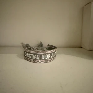 Grått armband från Christian Dior - Snyggt grått armband från Christian Dior med broderad logga i vitt. Armbandet har en bred, vävd design och avslutas med fransar. Perfekt accessoar för att lyfta din outfit med en exklusiv touch.