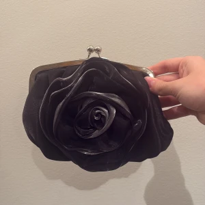 Svart kuvertväska med stor ros - Svart kuvertväska i glansigt tyg med en stor, dekorativ ros framtill. Väskan har silverfärgad metallram och klassiskt knäppspänne. Insidan har ett litet fack och en kedja för att bära väskan över axeln. Perfekt för att lyfta din outfit med en cool detalj.