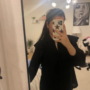 Svart klänning med volangärmar - Säljer en svart klänning med v-ringad hals och snygga volangdetaljer vid axlarna. Klänningen har långa ärmar med lätt puff och är i en loose passform. Perfekt för dig som gillar stilrena och trendiga plagg med lite extra attityd.