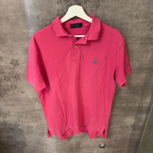 Rosa pikétröja från Polo Ralph Lauren - Snygg rosa pikétröja från Polo Ralph Lauren med klassisk krage och två knappar framtill. Tröjan har det ikoniska blå broderade logotypen på bröstet och är gjord i bomull. Perfekt för en chill och stilren look.
