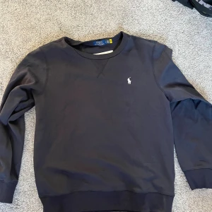 Mörkblå sweatshirt från Polo Ralph Lauren - Mörkblå sweatshirt från Polo Ralph Lauren med klassisk rund halsringning och broderad vit logga på bröstet. Tröjan har långa ärmar och ribbade muddar vid ärmslut och nederkant. Perfekt för en chill och stilren look.