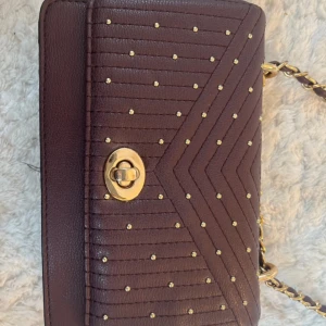 Axelväska med gulddetaljer - Snygg mörklila axelväska😍 Framsidan har quiltad design med små guldfärgade nitar i mönster. Insidan har fack för kort och dragkedja för småsaker. Säljer då den inte används längre. Liten slitning på bandet men inget man tänker på. Pris går att diskuteras💕