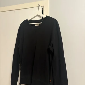 Svart sweatshirt från Nudie Jeans - En stilren svart sweatshirt från Nudie Jeans i 100% ekologisk bomull. Tröjan har rund halsringning, långa ärmar och en diskret logga nertill. Perfekt för dig som gillar enkel och clean streetstyle.