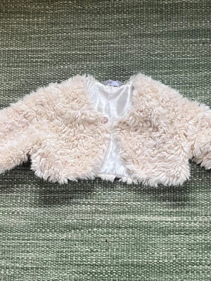 Beige fluffig bolero från Lindex Kids - Supersöt beige bolero från Lindex Kids med fluffig, lockig yta och glansigt innerfoder. Kort, croppad långärmad modell med en dekorativ knapp framtill. Perfekt för att lyfta vilken outfit som helst.
