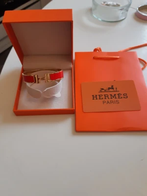Hermes armband  - Bilderna visar ett Hermès-armband i röd och guld färg med tillhörande ask och påse, 