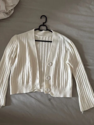 Vit ribbad kofta med stora knappar - Säljer en vit ribbad kofta med tre stora knappar framtill. Koftan är croppad och har lång ärm samt bred ribbad kant vid ärmslut och nederkant. Perfekt för lager på lager och enkel att styla till olika outfits.