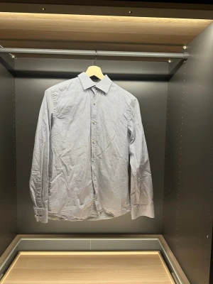 Vit och blå mönstrad skjorta Riley - Säljer en vit skjorta från Riley med små blå mönster och klassisk krage. Skjortan har lång ärm, knappar framtill och regular fit. Perfekt för dig som gillar stilrena detaljer och vill ha en skjorta som sticker ut lite.