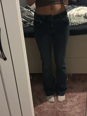 Mörkblå bootcut jeans med fickdetaljer - Säljer ett par mörkblå jeans med bootcut passform och snygga fickdetaljer bak. Jeansen har låg midja och klassisk femficksdesign. Materialet är jeans och de har dekorativa sömmar samt dubbla knappar i linningen.