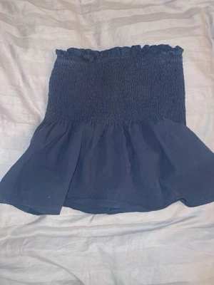 Mörkblå bandeau tubtopp med volang - Säljer en mörkblå bandeau tubtopp med smockad överdel och volang nedtill. Toppen är axelbandslös och har en stretchig passform som sitter snyggt på kroppen. Perfekt till sommaren och enkel att styla med jeans eller kjol.