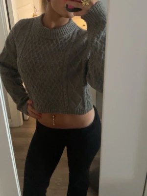 Grå croppad stickad tröja H&M - Säljer en grå croppad stickad tröja från H&M i storlek S. Tröjan har ett snyggt kabelstickat mönster, rund halsringning och långa ärmar. Perfekt för lager på lager och ger en chill vibe till din outfit.