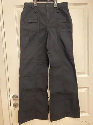 Svarta cargopants med bred passform - Säljer ett par svarta cargopants med breda ben och elastisk midja. Byxorna har stora fickor framtill med lock och bälteshällor. Tillverkade i ett slitstarkt bomullsmaterial och har en loose fit som ger en avslappnad look. Aldrig använda.