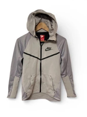 Nike Tech Fleece hoodie - L - 147-158 cm - Grå Nike Tech Fleece hoodie med dragkedja och huva. Jackan har två fickor fram och en svart Nike logga på bröstet. Detaljer i svart. Till barn mellan 12-13 år 