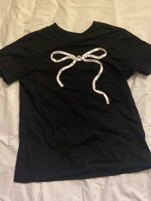 Svart t-shirt med rosettdetalj från Divided - Svart t-shirt från H&M Divided i storlek S. Framtill sitter en stor vit rosettdetalj i satinband som ger en söt och trendig look. Klassisk rund halsringning och korta ärmar. Supersöt med lite coquette vibe och är perfekt att ha med ett par jeans eller liknande. 😊