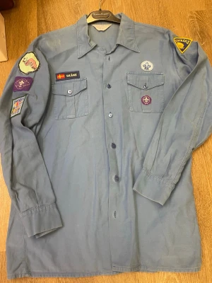 Blå scoutskjorta med märken - Blå långärmad scoutskjorta med flera broderade märken på ärmar och bröst, bland annat Skåne-flagga och scoutlilja. Skjortan har två bröstfickor med knappar och klassisk krage. Perfekt för dig som vill sticka ut på scoutäventyr.