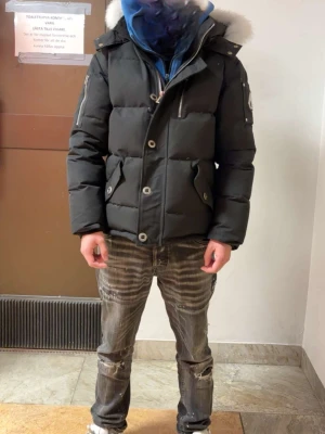 Svart Moose Knuckles dunjacka - Säljer en svart dunjacka från Moose Knuckles med fet puffer-look och vit päls runt huvan. Jackan har dragkedjor på ärmarna, stora knappar framtill och logga på armen. Perfekt för kalla dagar och riktigt snygg streetstil.