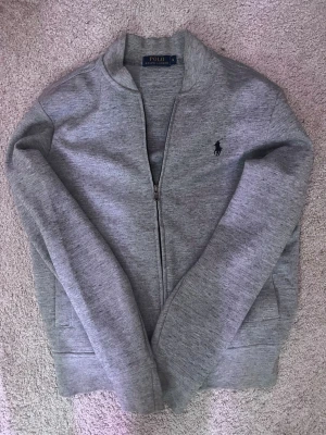 Grå ziptröja från Polo Ralph Lauren - Säljer en stilren grå ziptröja från Polo Ralph Lauren med diskret logga på bröstet. Tröjan har lång ärm, normal passform och dragkedja framtill. Perfekt till chill dagar och enkel att matcha med det mesta.